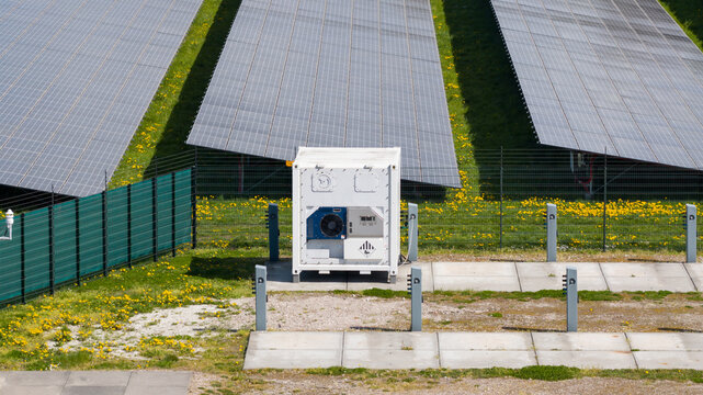 Aerial view of a solar farm featuring rows of photovoltaic panels and a white industrial inverter container set amidst green grass with yellow dandelions in Opmeer, North Holland, Netherlands.