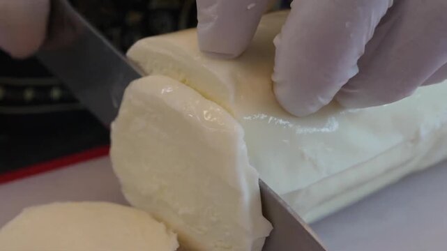 High-quality close-up footage of a chef wearing protective gloves slicing a log of fresh Mozzarella Filone cheese on a cutting board. This traditional low-moisture Italian cheese is a primary ingredie