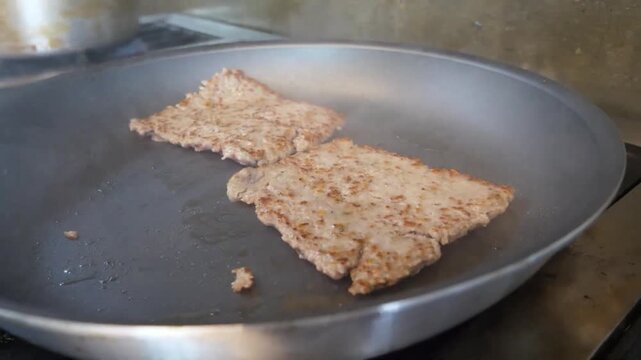 A gloved hand uses a wooden spatula to cook square-shaped raw minced pork or beef meat patties in a non-stick frying pan, preparing fresh toppings for a traditional homemade Italian pizza.