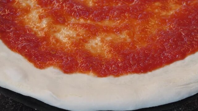 A close-up shot capturing the process of making traditional Italian pizza, showing vibrant red tomato sauce being spread in a circular motion over fresh, white raw dough on a floured surface. This foo