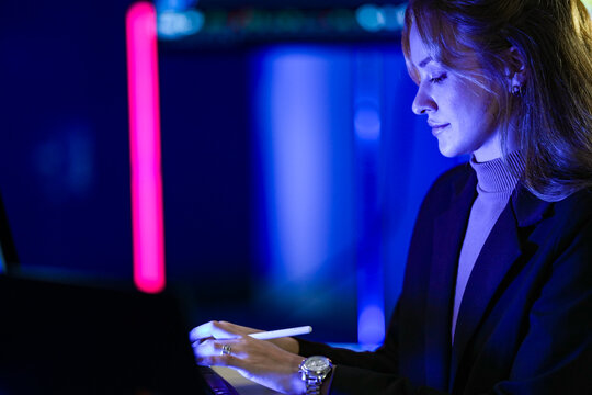 Young businesswoman with long fair hair working with a stylus at monitors of program code marks a bug with the stylus tip while the team records a technology explainer in the darkened