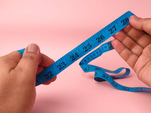 Two hands hold and unroll a coiled blue plastic measuring tape displaying numbers against a pink background