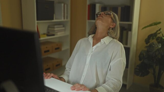 Woman clutching hands to chest at office desk with computer and bookshelf, leaning back with eyes closed and glasses tilted; heartattack anxiety.