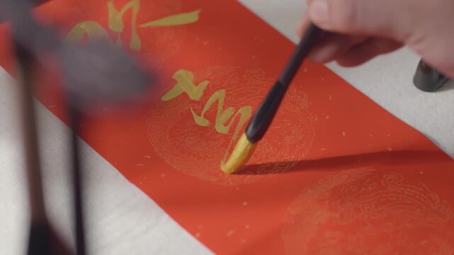 Senior man artist writes the chinese calligraphy using golden ink on crimson paper, skillful brush strokes create elegant characters with wisdom writing at a wooden desk on red paper