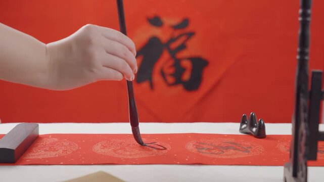 Artist hand practicing writes traditional chinese calligraphy using golden ink on crimson paper, skillful brush strokes create elegant characters with wisdom writing at a wooden desk on red paper