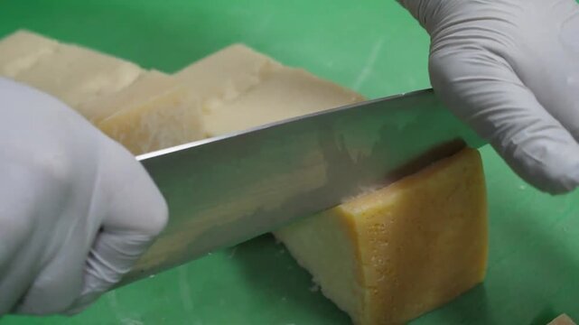 Close-up of a chef's hands in white gloves using a knife to cut a block of aged Italian cheese, such as Parmigiano Reggiano or Grana Padano, on a green cutting board in a professional kitchen. Perfect