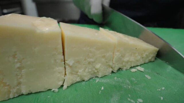 A professional chef wearing white gloves carefully cuts a large wedge of aged Italian hard cheese, such as Parmigiano-Reggiano or Grana Padano, on a green commercial cutting board. This close-up foota