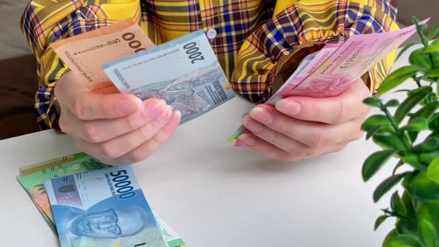 A close-up of a woman counting Indonesian rupiah banknotes at a table. Concepts for personal finance, budgeting, cash payments, travel, and money transactions.