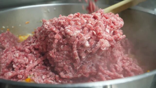 High-angle close-up of a chef in a professional kitchen adding raw minced beef to a large stainless steel pot containing saut&eacute;ed carrots, onions, and celery (soffritto). This footage demonstrates the 