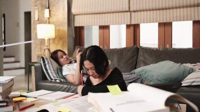 Two Asian teenage tutorials, girl concentrates on learning class at home with pile of homework, her friend uses mobile phone on sofa, self-study with textbooks, homeschooling together lifestyle.