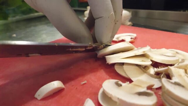 A professional chef uses a sharp knife to carefully slice fresh white champignon mushrooms on a red cutting board. This high-quality footage captures the detailed preparation of organic ingredients fo