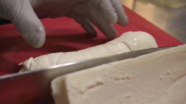 Close-up shot of a professional chef's hands meticulously slicing a fresh, soft mozzarella cheese log on a red cutting board with a sharp stainless steel knife. Perfect for culinary concepts, Italian 