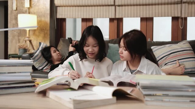Two Asian teenage tutorials, girl concentrates on learning class at home with pile of homework, her friend uses mobile phone on sofa, self-study with textbooks, homeschooling together lifestyle.