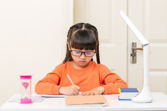 Focused Asian schoolgirl doing homework with hourglass timer, time management and study discipline concept