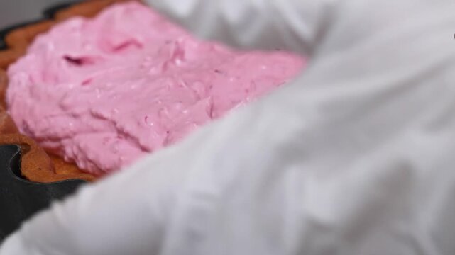 close-up shot of a chef or baker using a spatula to spread a light, fluffy pink berry cream or mousse filling into a pre-baked golden fluted tart shell. This vibrant summer dessert is perfect for culi