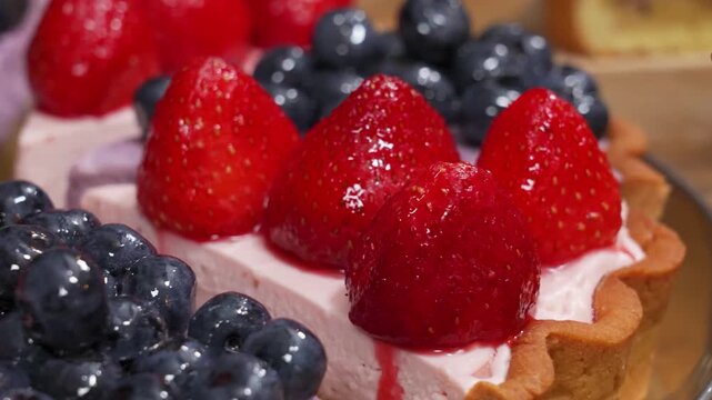 High-quality close-up footage of gourmet dessert slices featuring a creamy cheesecake filling on a golden crust. The slices are generously garnished with fresh, glossy blueberries and vibrant red stra