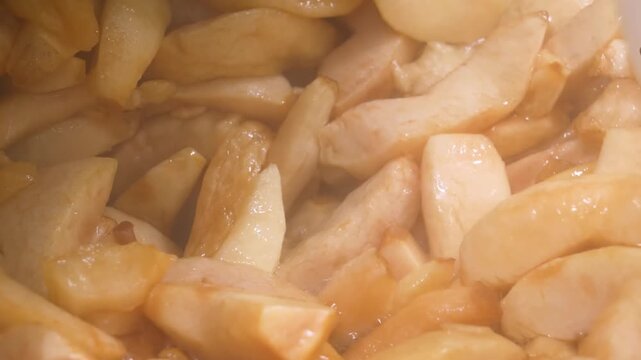 A detailed close-up shot of sliced fresh apples being cooked with cinnamon, brown sugar, and butter. The fruit is tender and coated in a thick, bubbling caramel sauce, perfect for homemade apple pie, 