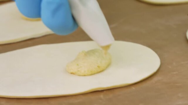 A professional baker wearing blue hygiene gloves uses a piping bag to apply a thick, creamy filling resembling almond cream or frangipane onto a circular sheet of raw pastry dough on a parchment-lined