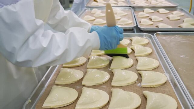 Close-up footage of a professional pastry chef preparing trays of raw dessert hand pies in a commercial kitchen. The scene shows multiple metal trays filled with uniform, crescent-shaped pastries bein