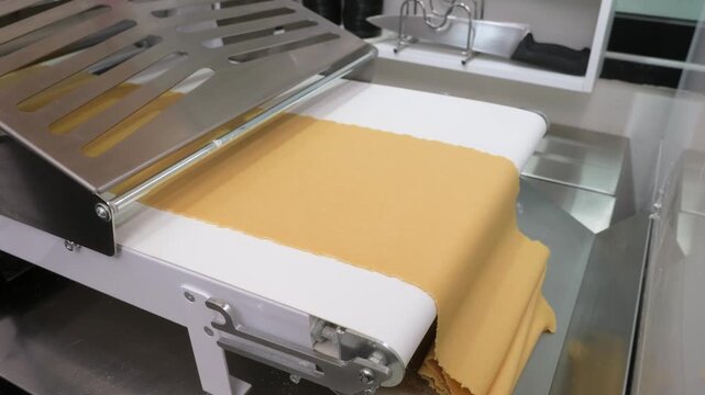 High-angle footage of an automatic dough sheeter at work in a modern commercial kitchen. The machine precisely rolls out smooth, thin layers of pastry dough on a white conveyor belt, highlighting the 
