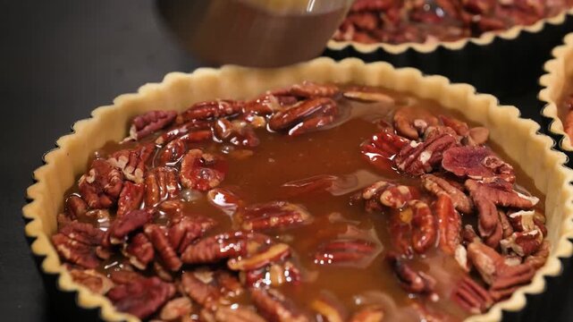 close-up, slow-motion shot showing luscious, golden caramel sauce being poured from a metal measuring cup onto a bed of toasted pecan halves inside a homemade fluted tart crust. Perfect for holiday ba