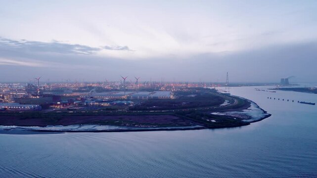 Drone footage of Antwerp Port during the evening, showing illuminated warehouses, the Scheldt River, wind turbines, and an open evening sky above the harbor landscape.