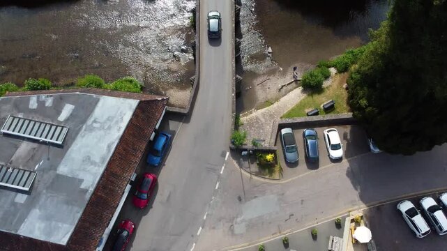 Small English village cottages, riverside parking and a tight road junction viewed from above on a bright summer day.