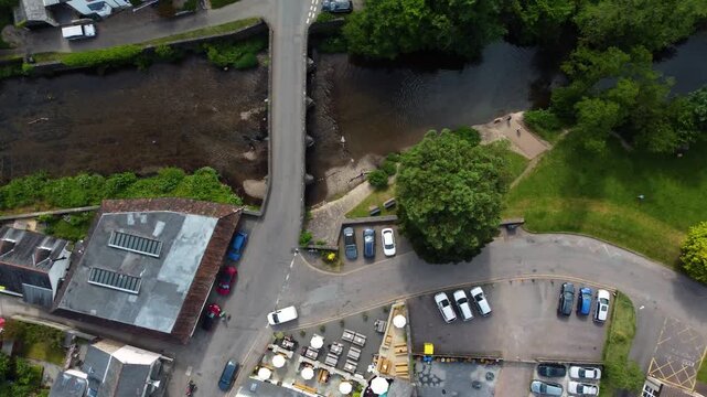 Cars move along a narrow rural road crossing a river beside a car park, trees and small commercial buildings in Exmoor, Devon.