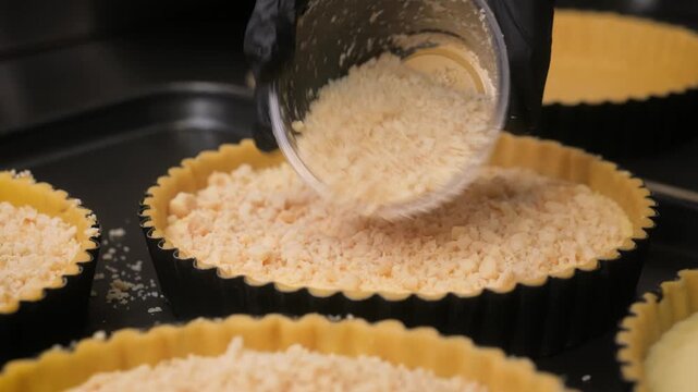 Close-up of a pastry chef&rsquo;s hands in black gloves carefully sprinkling golden biscuit crumble onto a creamy yellow custard tart filling in a fluted black baking tin. Part of a professional bakery prod