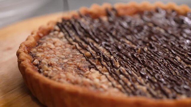 Close-up footage of a decadent homemade dessert pie featuring a golden fluted crust and a rich nut filling (likely pecan or hazelnut), being finished with a artistic drizzle of smooth, glossy dark cho