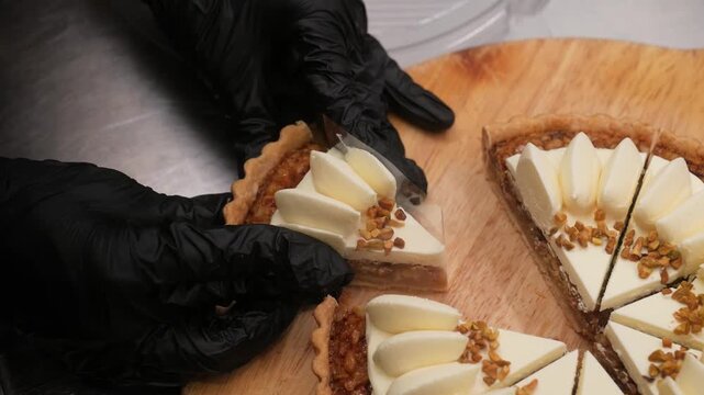 Close-up of a pastry chef wearing black gloves carefully arranging slices of an artisanal nut tart on a wooden board. The dessert features a buttery shortcrust base, rich caramel or pecan cream fillin