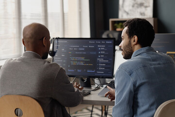 Medium shot of two middle aged men reviewing software code on desktop monitor in information technology office, collaborating on debugging and team workflow. For IT services marketing © Seventyfour