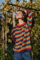Fototapeta premium Woman in a colorful striped sweater outdoors among vines, relaxed and smiling with short hair, casual outfit, hand behind head, warm golden hour lifestyle portrait.