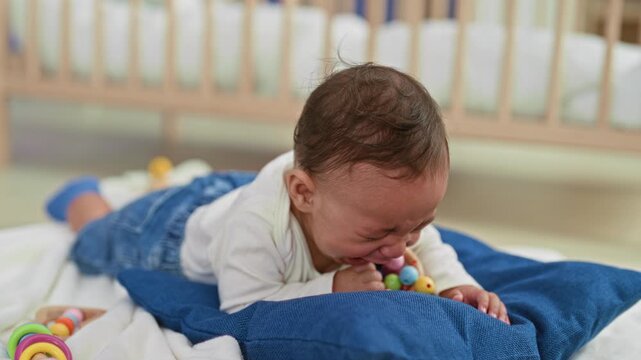 Baby boy crying with mouth open holding colorful rattle on blue pillow on bed in building; sadness vulnerability.