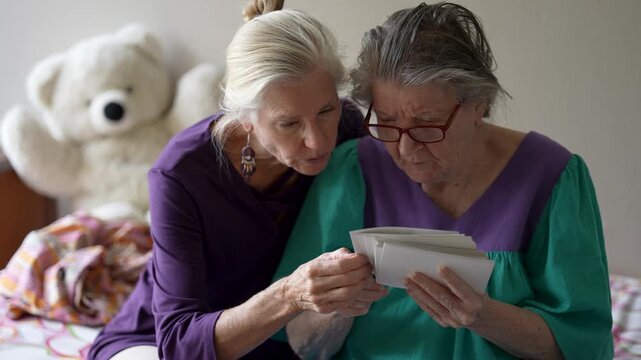 A mature daughter visits her elderly mother in a senior care facility. They share photos and stories while spending time together.
