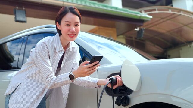 Smiling Asian woman using a smartphone while plugging a charger cable into an electric car at home. Concept of smart EV application and green technology.