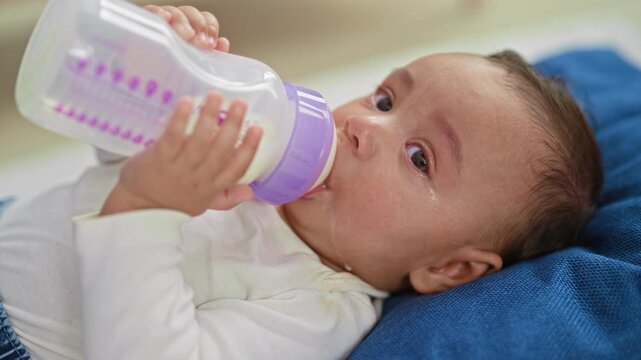 Hispanic baby boy holds bottle to mouth while feeding milk, drool and tears visible as he lies on a blue pillow in a building; comfort bonding.