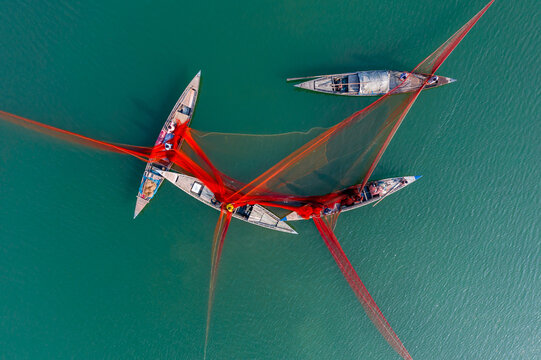 Bogura, Rajshahi Division, Bangladesh - 08 February 2024: Aerial view of fishermen on wooden boats casting large orange fishing nets in the Jamuna River.