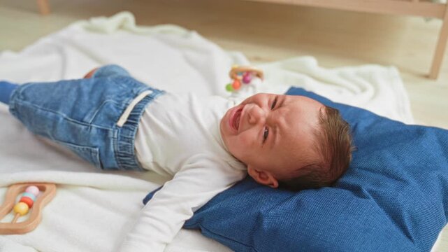 Toddler boy crying with tear and outstretched arm on blue pillow beside wooden rattle on blanket in building; distress seeking comfort.