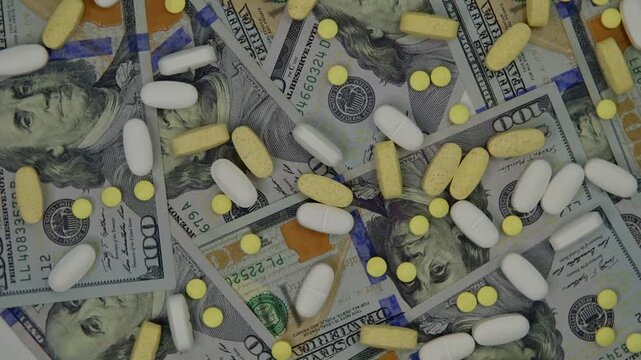 Various prescription drugs spilled out over a hundred-dollar bill, rotating right to left, viewed from overhead.