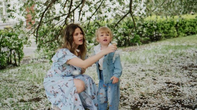 Mother gently guides toddler under blooming tree. Soft light filters through spring leaves. Child looks up with wonder and trust. Mother smiles warmly, supporting child's exploration