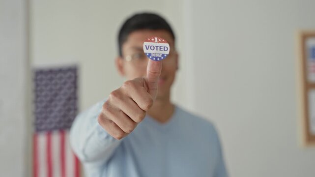 Man in light blue shirt points finger to a voted sticker on his thumb in a polling building with an american flag visible; civic pride.