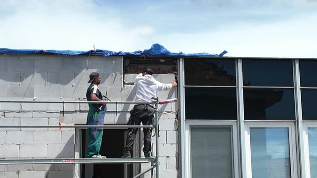 Construction workers on scaffolding installing windows on a building facade. Two men in work clothes are on a scaffolding platform, actively engaged in the process of installing windows on a building