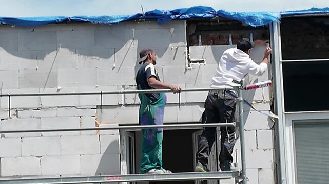 Construction workers installing windows on a brick building facade using scaffolding. Workers on scaffolding are sealing window frames and applying sealant to the exterior of a building under