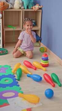 Girl kneeling and smiling, touching finger to lips beside colorful toy pins, ball and stacking rings on a wooden floor in a playroom building; playful joy.