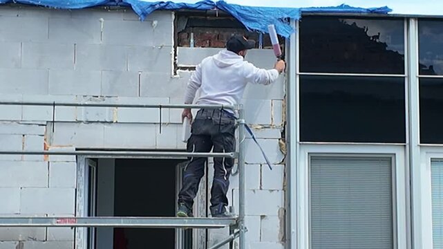 Construction worker installing windows on scaffolding at a building site. A worker in protective gear uses tools to install window frames on a building under construction, seen from scaffolding.