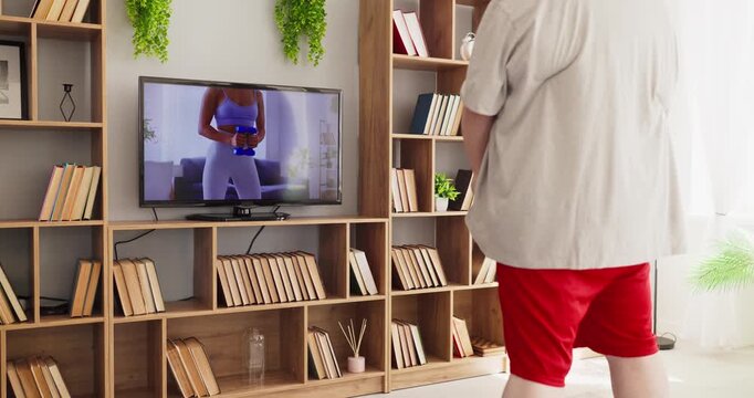 Overweight man home workout online fitness exercise. He follows a training class on TV, copying dumbbell moves in a living room with focus for weightloss. Concept of wellness and motivation.
