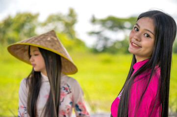 Fototapeta premium Two young beauty Vietnamese girls in traditional Ao Dai costumes and conical hat enjoying in green nature rice field 