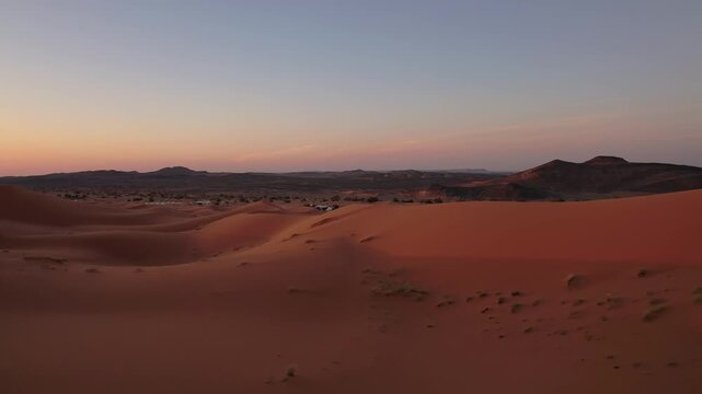Erg Chebbi dune sea near Merzouga, Morocco, reveals rolling sand formations, a luxury desert camp and distant rocky ridges illuminated by warm sunrise light, drone forward flight shot.