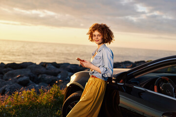 Fototapeta premium Cheerful woman outdoors by a car at sunset, wearing a denim shirt and mustard skirt, enjoying a relaxed lifestyle with coastal breeze and warm tones and golden light on her smile